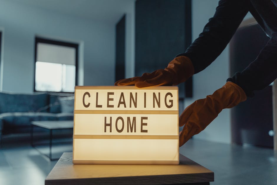 A person wearing brown cleaning gloves places a lightbox sign with the words 'CLEANING HOME' in black capital letters on a wooden table inside a modern living room. The room features a dark-colored so